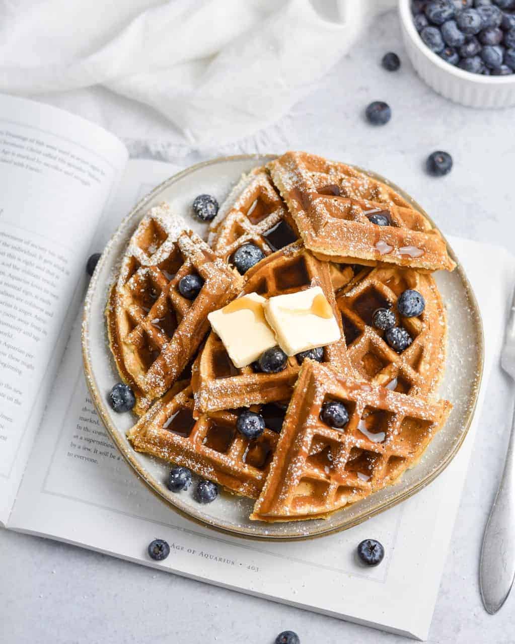 a stack of vegan belgian waffles on a plate topped with vegan butter and maple syrup. 