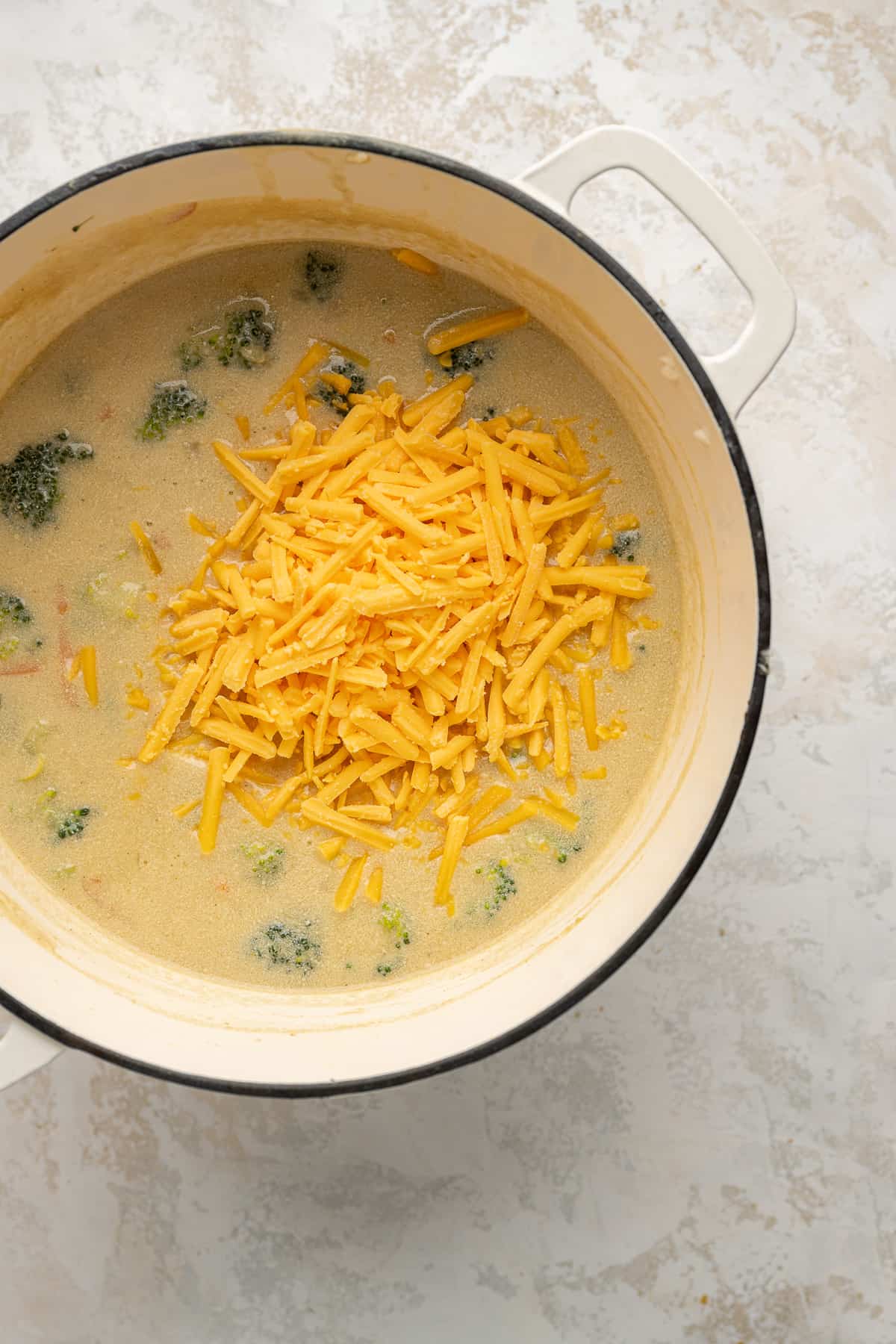 cheddar shreds being added to broccoli cheddar soup in a white dutch oven.